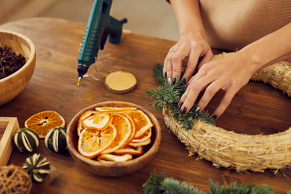 Close-up of unrecognizable woman with manicure standing at wooden table in workshop and using hot glue while attaching fir tree twigs to wreath base Photograph by Mediaphotos Close-up of unrecognizable woman with manicure standing at wooden table in workshop and using hot glue while attaching fir tree twigs to wreath base Photograph by Mediaphotos