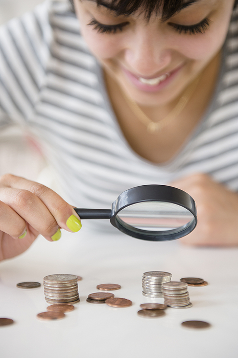 Mixed race woman examining stacks of coins with magnifying glass Photograph by JGI/Jamie Grill