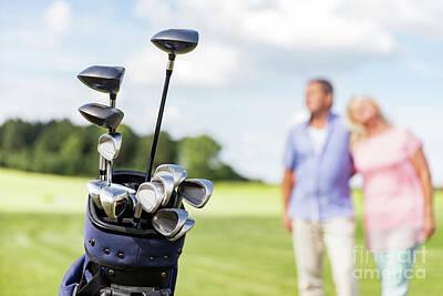 Sports Photos - Set of golf clubs with senior couple in the background. by Michal Bednarek